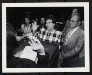 Black-and-white photograph of Esther Campos being sworn in, possibly as a new Houston Independent School District (HISD) board member. She is accompanied by her husband, Jesus Campos.