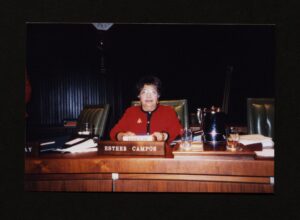 Color photograph of Esther Campos in red suit seated at her assigned chair on the Houston Independent School District (HISD) Board of Education Trustee dais.